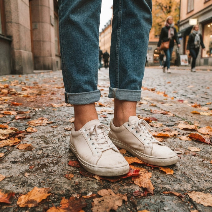 Vita sneakers och uppvikta jeans på höstlöv på en trottoar
